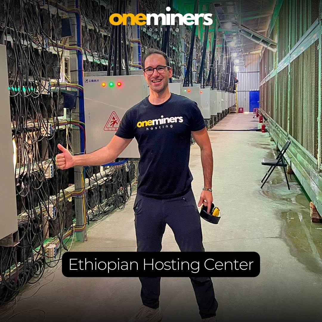 CEO of OneMiners posing in the Ethiopian hosting center, with rows of mining equipment in the background and a 'OneMiners Hosting' t-shirt.